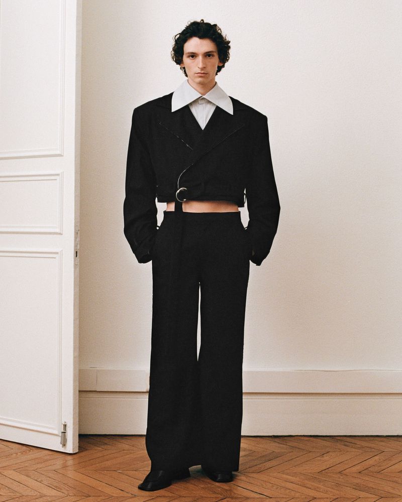 Male fashion model in a black cropped suit jacket and wide-leg trousers standing in a simple interior with wooden floor and white door.
