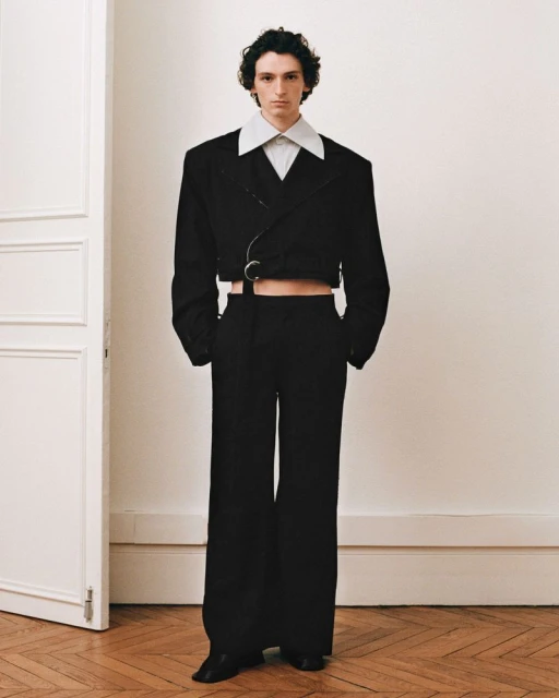 Male fashion model in a black cropped suit jacket and wide-leg trousers standing in a simple interior with wooden floor and white door.