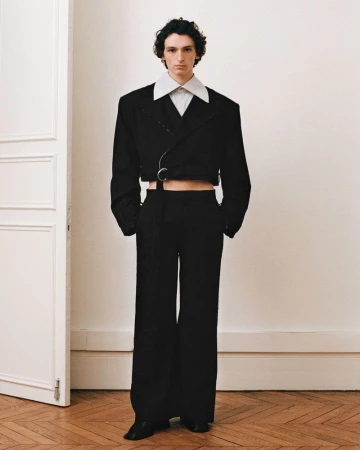 Male fashion model in a black cropped suit jacket and wide-leg trousers standing in a simple interior with wooden floor and white door.