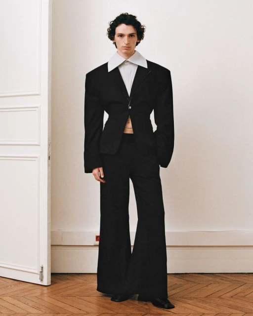 A slender man posing in a black suit with wide-leg pants and an oversized jacket stands against a plain wall in a fashion-style image.