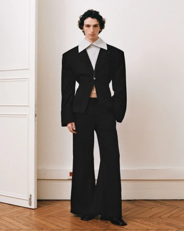 A slender man posing in a black suit with wide-leg pants and an oversized jacket stands against a plain wall in a fashion-style image.