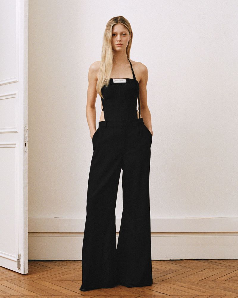 Adult woman poses in a black halter jumpsuit in an indoor photo studio with a white wall, herringbone wood floors, and a white door.