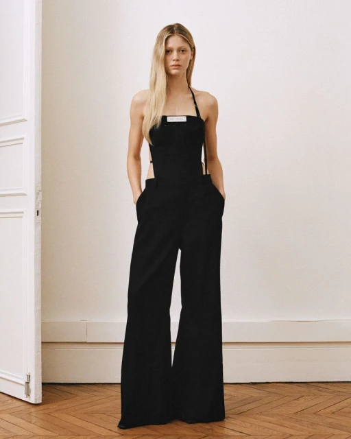Adult woman poses in a black halter jumpsuit in an indoor photo studio with a white wall, herringbone wood floors, and a white door.