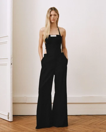 Adult woman poses in a black halter jumpsuit in an indoor photo studio with a white wall, herringbone wood floors, and a white door.