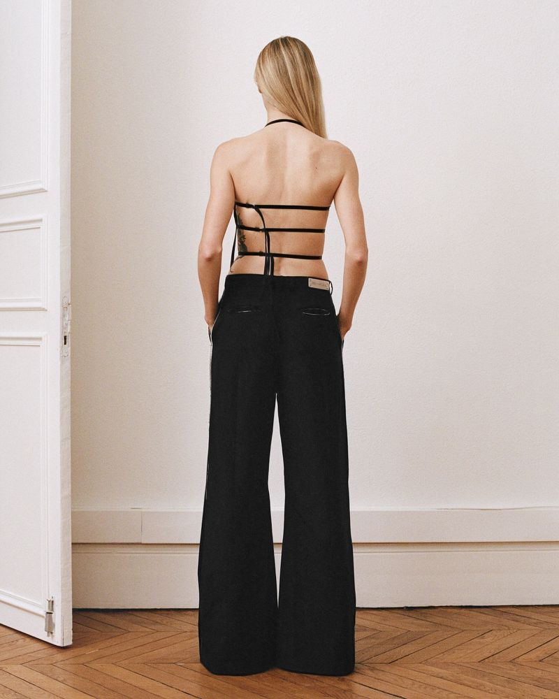 Back view of a blonde woman wearing a black strappy top and wide-leg pants, posing against a white wall and wooden floor backdrop.