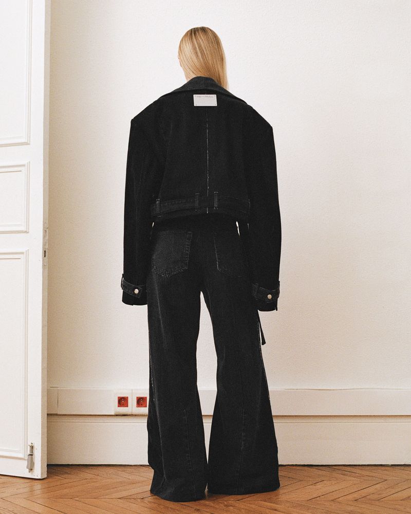 Woman in oversized black denim jacket and wide-leg jeans stands facing away against a white wall, minimalist fashion aesthetic.