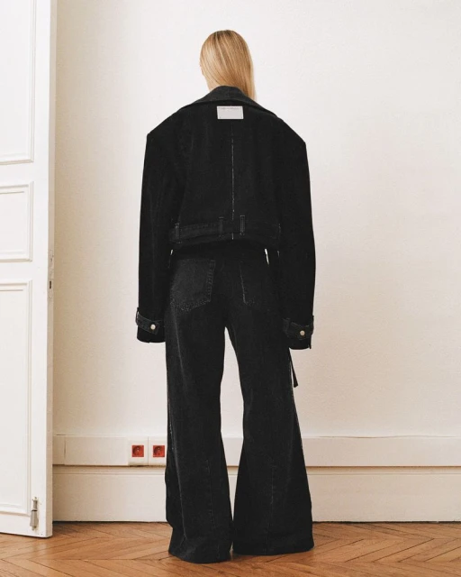 Woman in oversized black denim jacket and wide-leg jeans stands facing away against a white wall, minimalist fashion aesthetic.