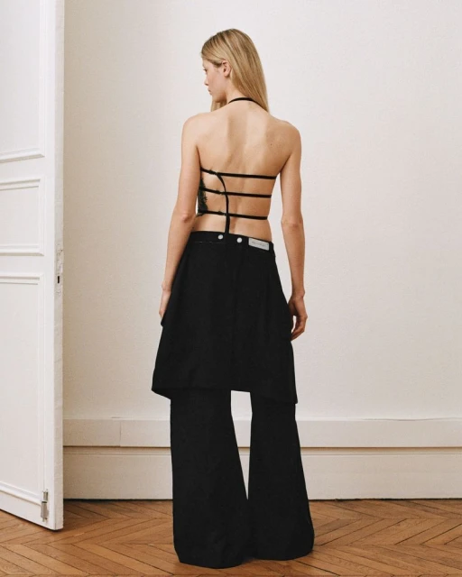 Back view of a slender blonde woman posing indoors, wearing black top and draped black pants in a minimalist setting with neutral tones.