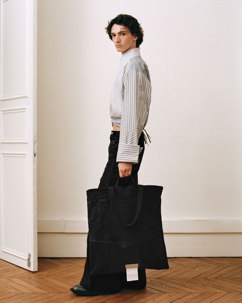 Fashionable male model showcases denim tote bag in a light studio, wearing pinstripe shirt and black wide-leg jeans, captured in a profile shot.
