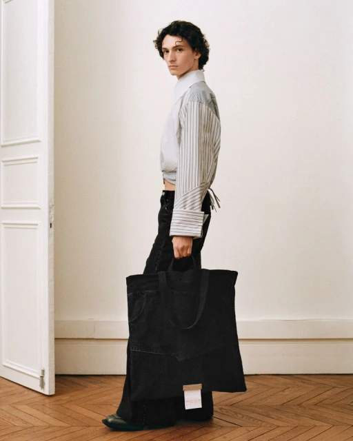 Fashionable male model showcases denim tote bag in a light studio, wearing pinstripe shirt and black wide-leg jeans, captured in a profile shot.