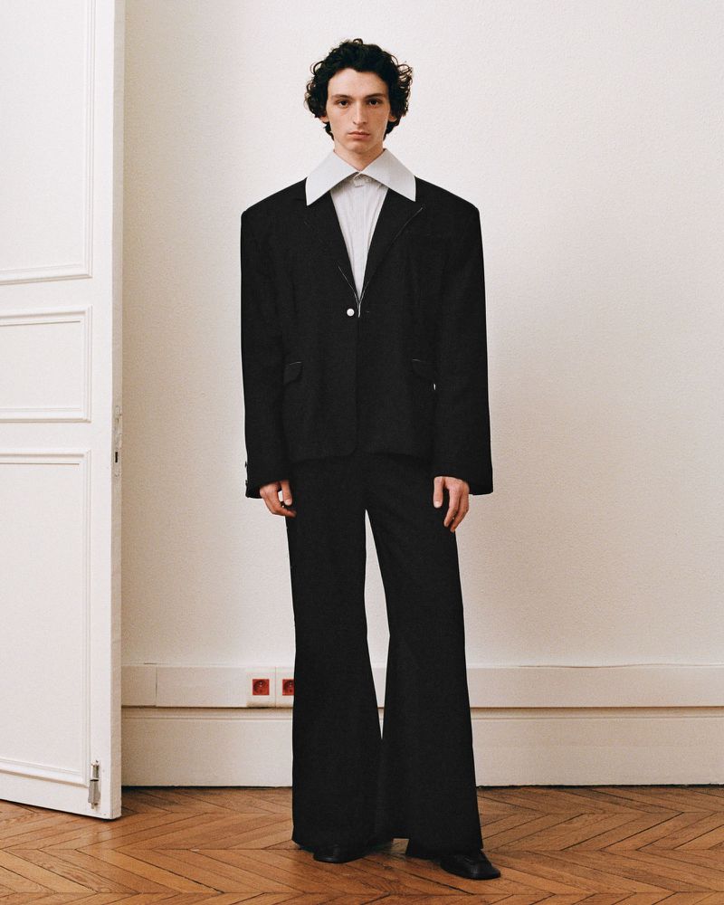 Full-length portrait of a male model wearing a black blazer with an oversized white collar and black trousers against a neutral interior wall.
