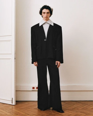Full-length portrait of a male model wearing a black blazer with an oversized white collar and black trousers against a neutral interior wall.