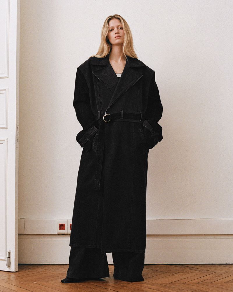 A tall, blond woman stands in a photo studio wearing a dark denim trench coat with a belt over dark denim wide-leg pants.