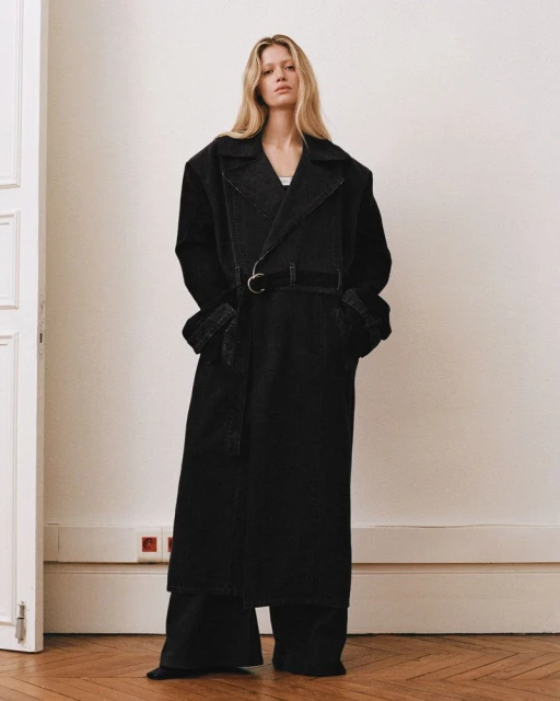 A tall, blond woman stands in a photo studio wearing a dark denim trench coat with a belt over dark denim wide-leg pants.