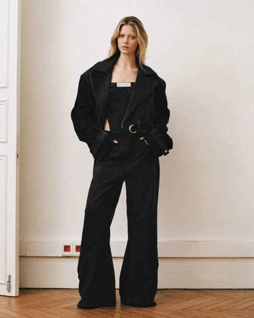 Full-length shot of a blonde female model wearing a black denim trench coat and bell-bottom jumpsuit in front of a white wall.