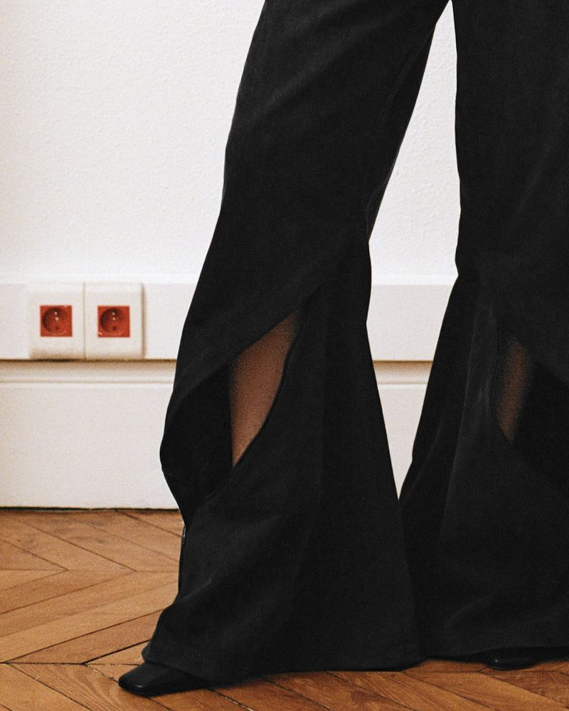 Fashionable black wide-leg pants with side slits and pointed shoes against a white wall and wood flooring.
