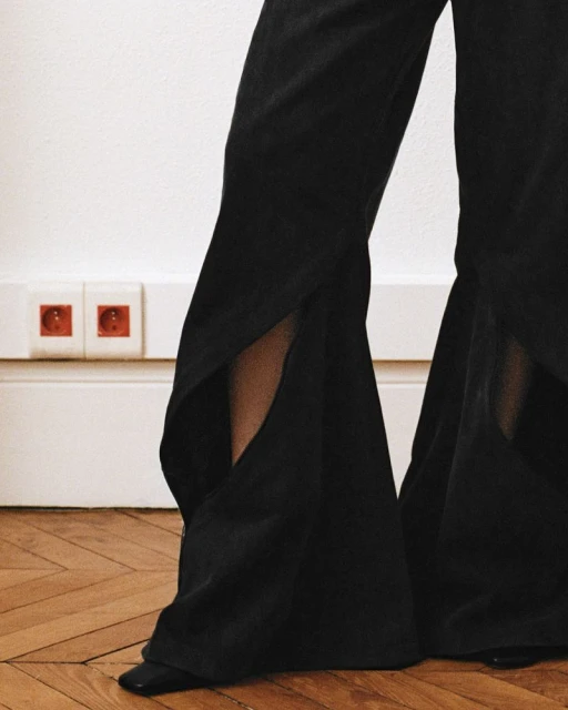 Fashionable black wide-leg pants with side slits and pointed shoes against a white wall and wood flooring.