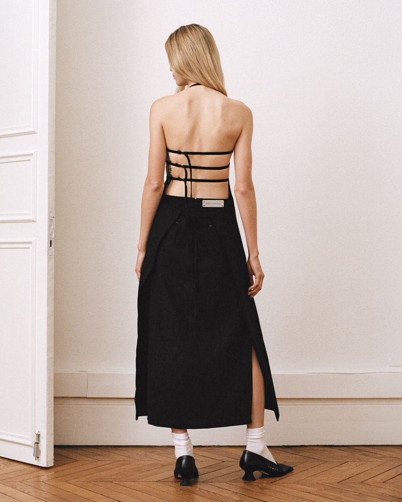 Back view of a blonde woman in a black halter top and skirt with slits, standing on a hardwood floor in front of a white wall.