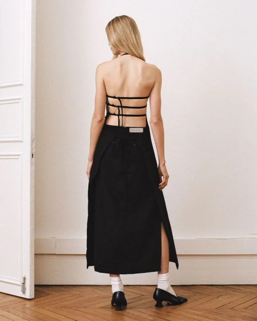 Back view of a blonde woman in a black halter top and skirt with slits, standing on a hardwood floor in front of a white wall.