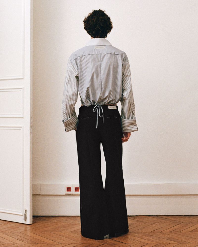 Back view of an adult in a striped collared shirt and black wide-leg trousers, standing indoors against a neutral wall and doorway.