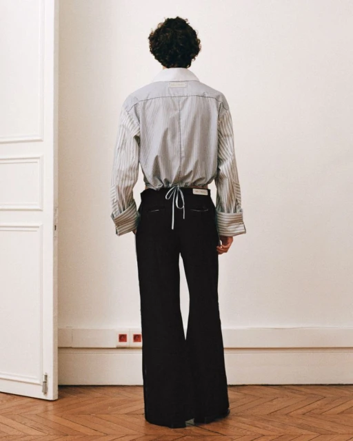Back view of an adult in a striped collared shirt and black wide-leg trousers, standing indoors against a neutral wall and doorway.