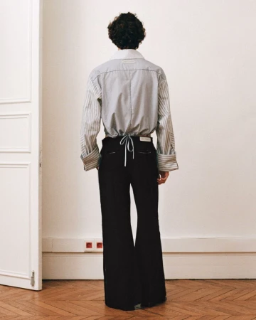 Back view of an adult in a striped collared shirt and black wide-leg trousers, standing indoors against a neutral wall and doorway.