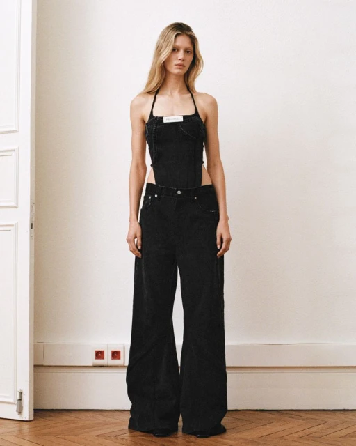A blonde fashion model poses in an indoor studio wearing a black denim halter top and baggy denim pants, showcasing a modern, edgy style.