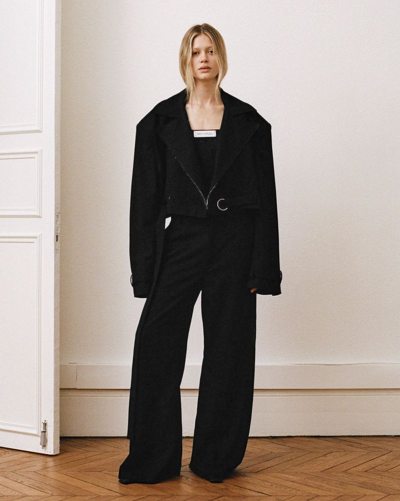 Model standing in a black blazer and matching trousers in a light interior with herringbone parquet flooring.