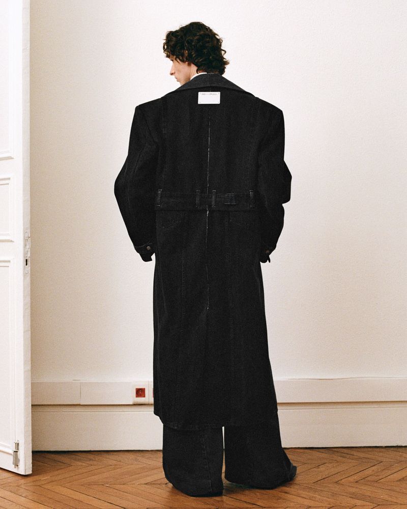 Model with dark wavy hair wearing an oversized black coat and matching trousers shot from behind in an interior space