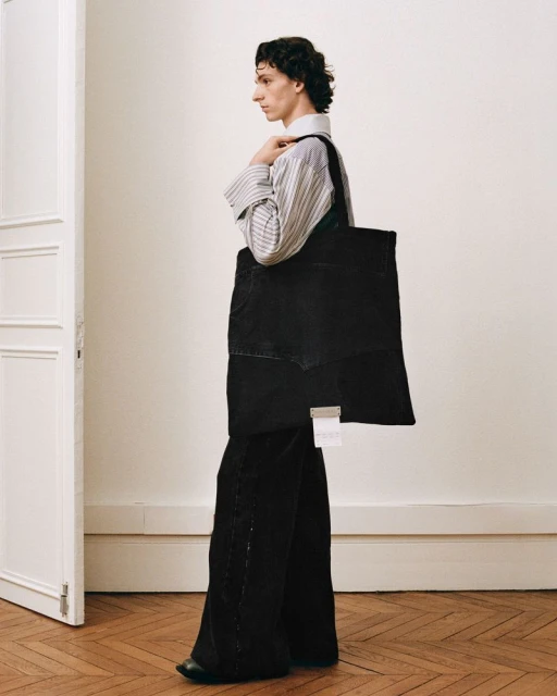 Side view of a stylish adult wearing a striped shirt, wide black pants, and carrying a black tote bag in an indoor setting.