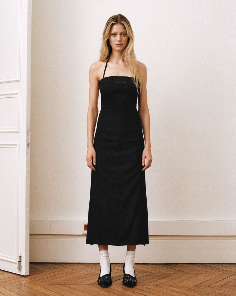 Full-length studio shot of a blonde woman modeling a black halter dress with a thigh slit, paired with white socks and black shoes.