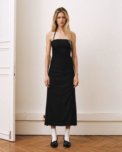 Full-length studio shot of a blonde woman modeling a black halter dress with a thigh slit, paired with white socks and black shoes.