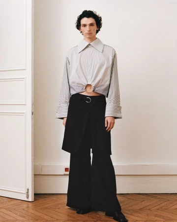 Model standing in a room wearing a cropped striped shirt with a metal ring cutout and wide black trousers.