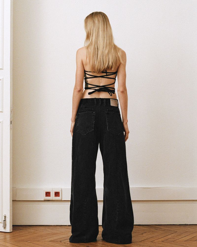 Back view of a blonde adult female wearing a black criss-cross top and wide-leg black jeans, standing indoors in a minimalistic room.