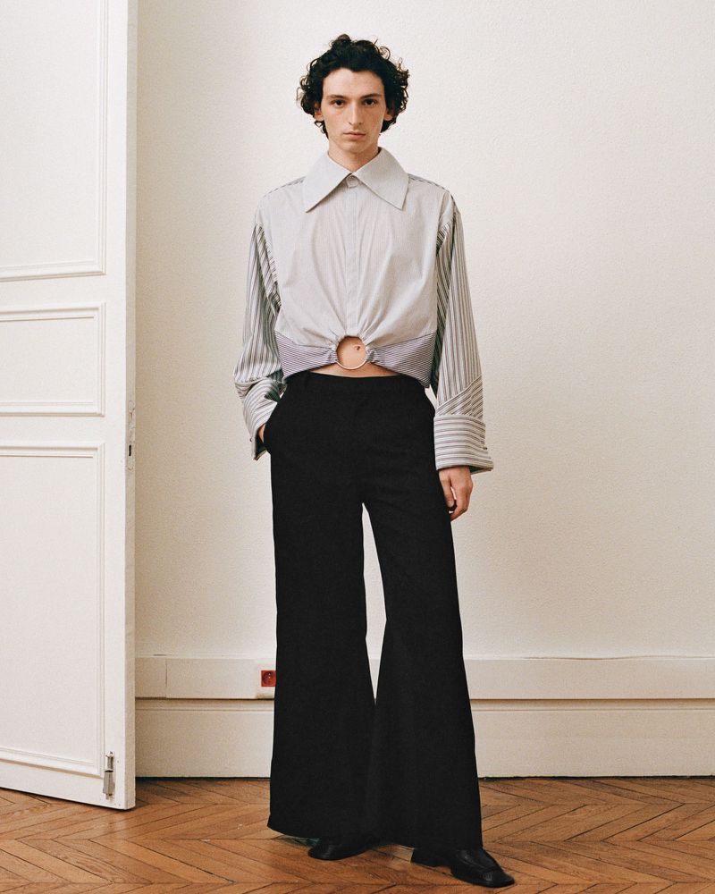 Fashionable adult posing indoors wearing a striped long-sleeve shirt with an o-ring detail and black wide-leg pants in a fashion editorial.