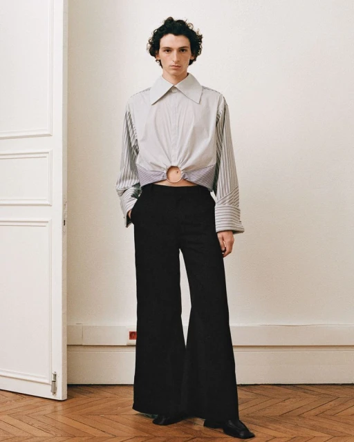 Fashionable adult posing indoors wearing a striped long-sleeve shirt with an o-ring detail and black wide-leg pants in a fashion editorial.