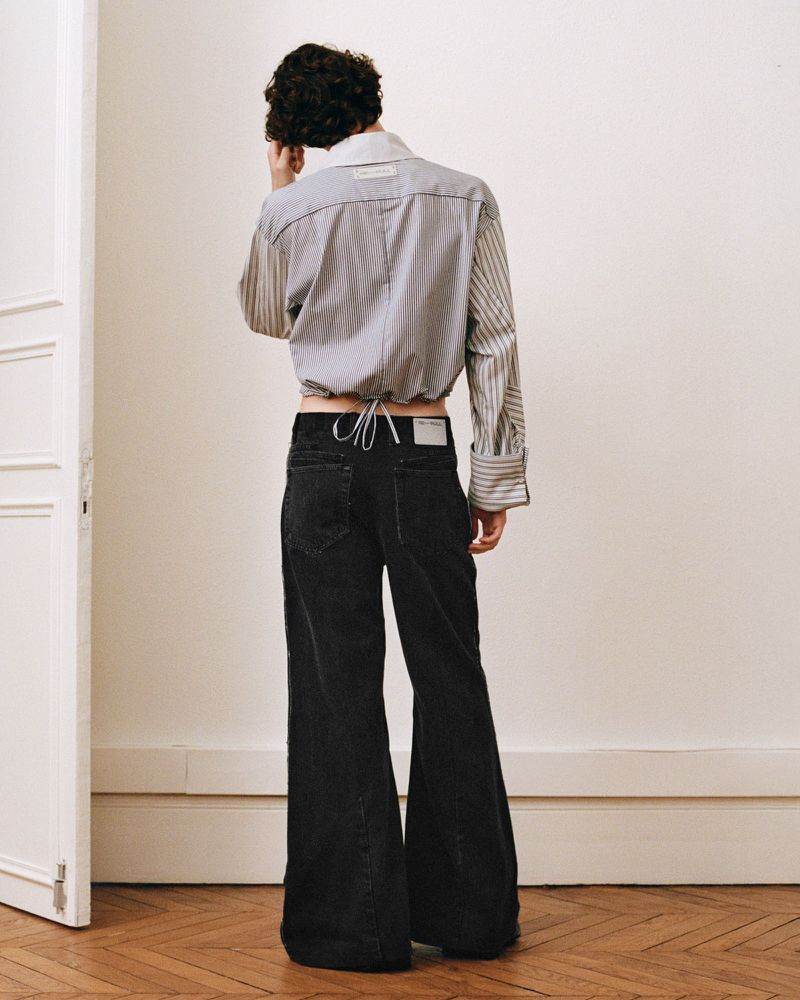Back view of a person wearing a striped crop top and black bell-bottom jeans standing on a wooden floor in front of a white wall.
