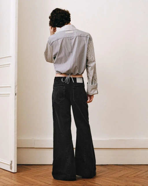 Back view of a person wearing a striped crop top and black bell-bottom jeans standing on a wooden floor in front of a white wall.