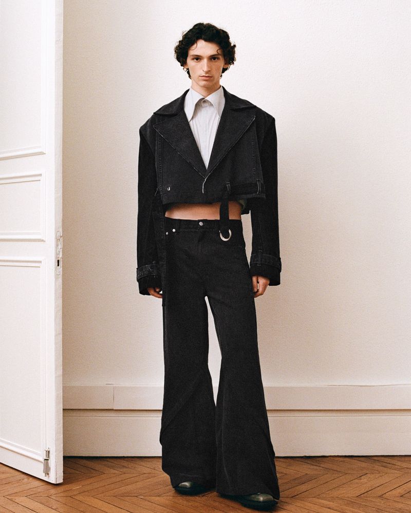 Male model in a black cropped denim jacket and wide-leg pants poses in front of a neutral wall on a wooden floor in a fashion editorial.