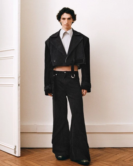 Male model in a black cropped denim jacket and wide-leg pants poses in front of a neutral wall on a wooden floor in a fashion editorial.