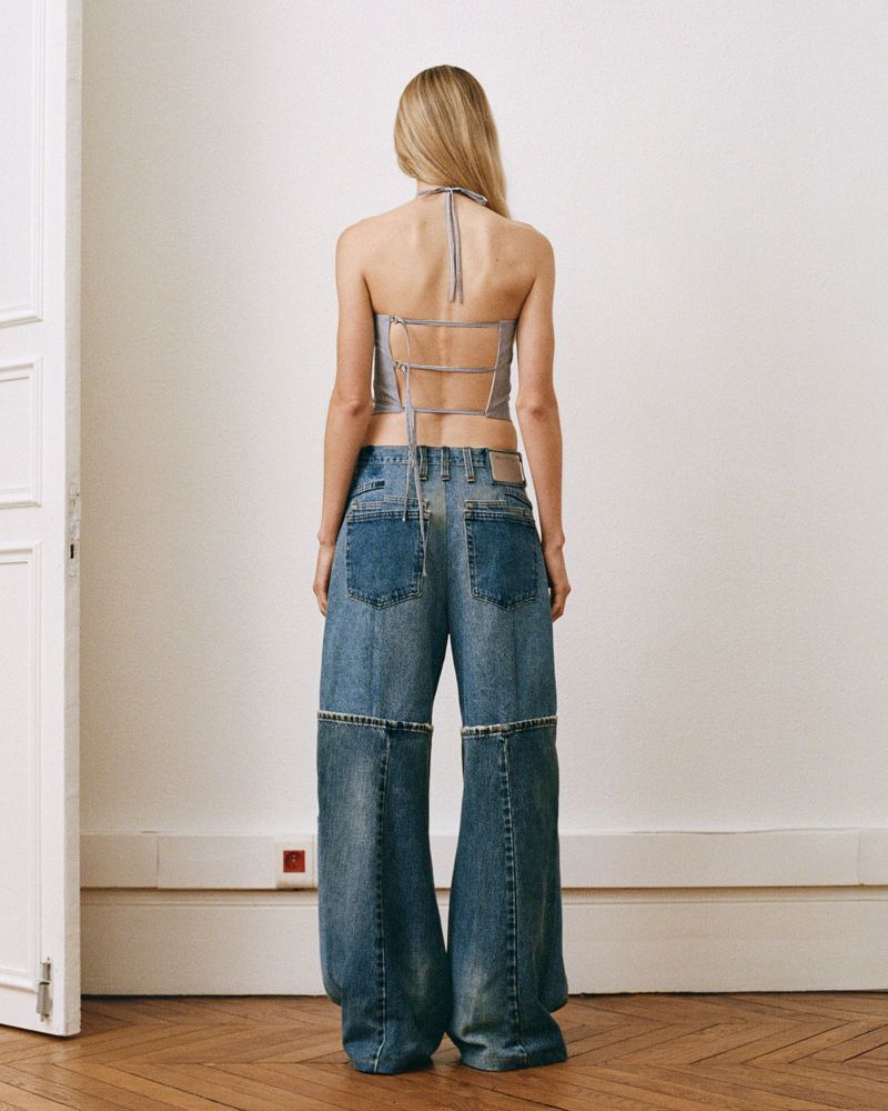 A woman stands with her back to the camera wearing a grey top and wide-legged jeans, posing indoors with a white wall and wooden floor.