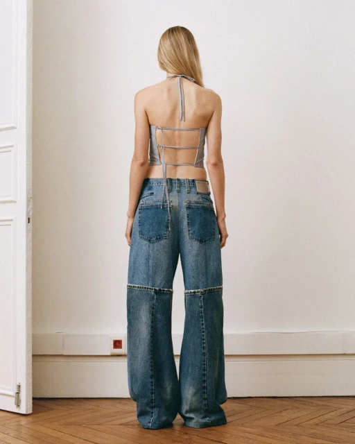 A woman stands with her back to the camera wearing a grey top and wide-legged jeans, posing indoors with a white wall and wooden floor.