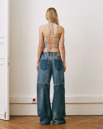 A woman stands with her back to the camera wearing a grey top and wide-legged jeans, posing indoors with a white wall and wooden floor.
