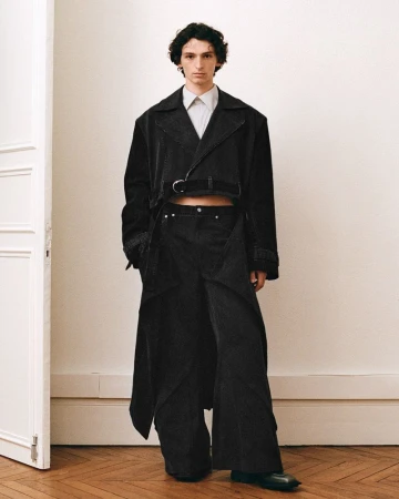 Man wearing a cropped black jacket and wide trousers standing in a room with a white door and wood parquet floor.