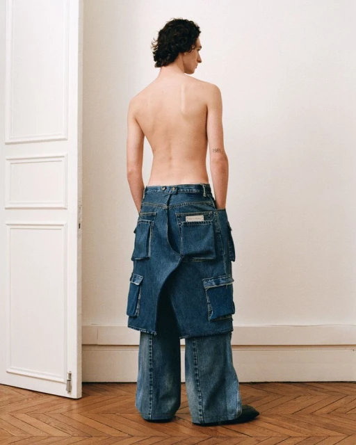 Back view of a shirtless male model wearing wide-leg patchwork blue jeans with pockets on a light parquet floor near a white door.