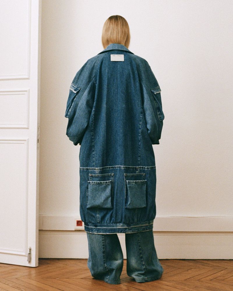 Back view of a woman in a long denim coat and wide-leg jeans, showcasing oversized fashion with a focus on textures and materials indoors.