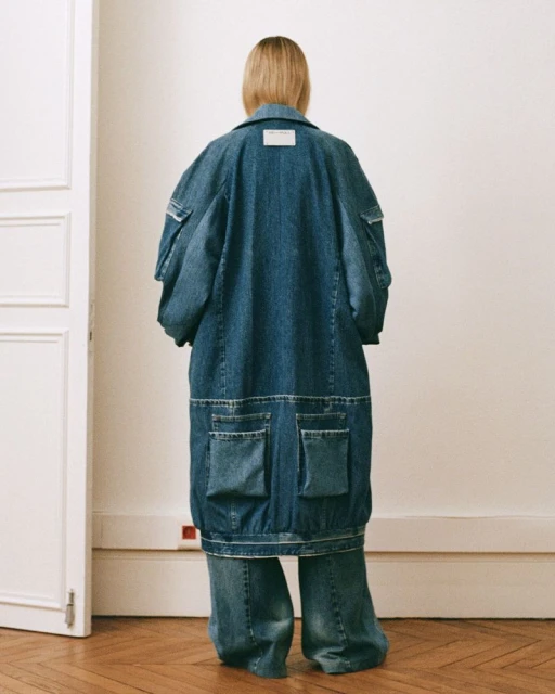 Back view of a woman in a long denim coat and wide-leg jeans, showcasing oversized fashion with a focus on textures and materials indoors.