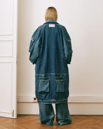 Back view of a woman in a long denim coat and wide-leg jeans, showcasing oversized fashion with a focus on textures and materials indoors.