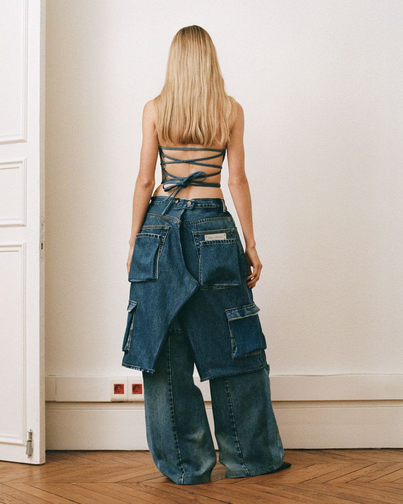 A woman with long blond hair is seen from the back, wearing a denim top and pants with layers of denim pockets. Modern fashion.