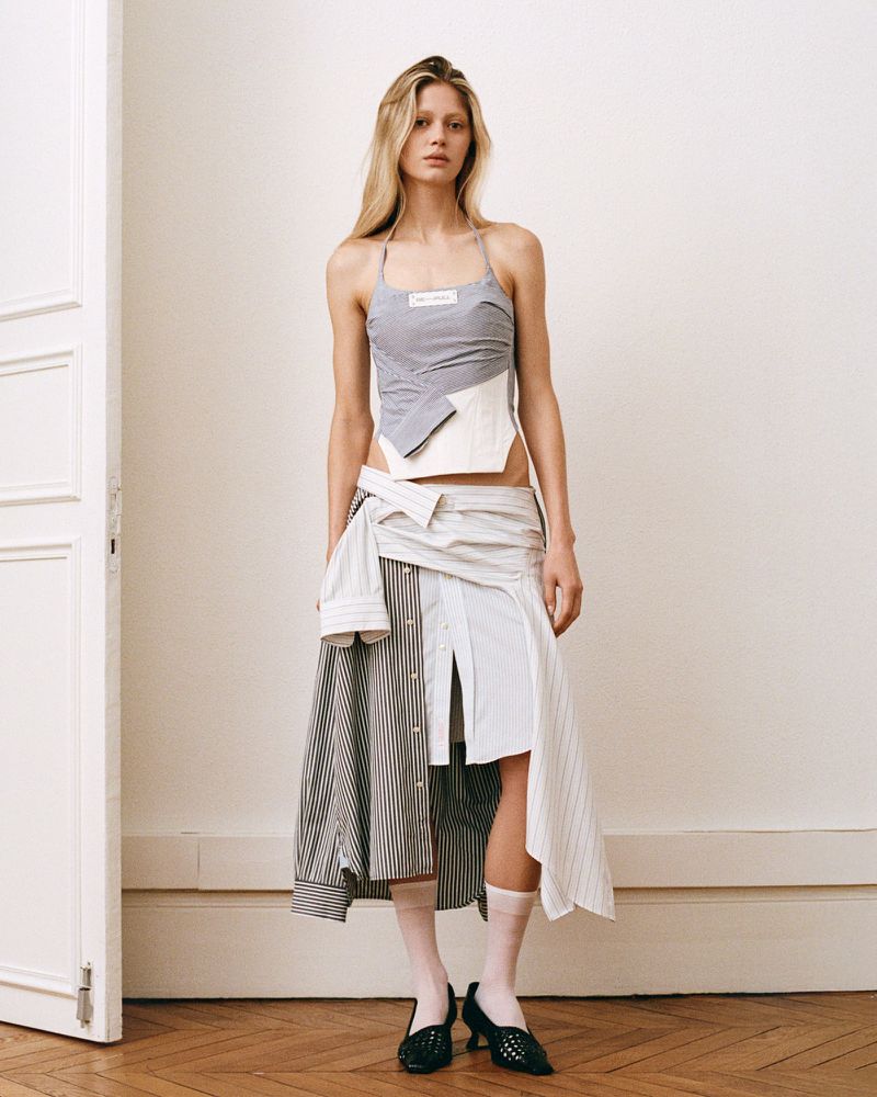 A fashionable adult female in a unique grey halter top and mixed-fabric skirt stands against an off-white wall with a partially visible white door.